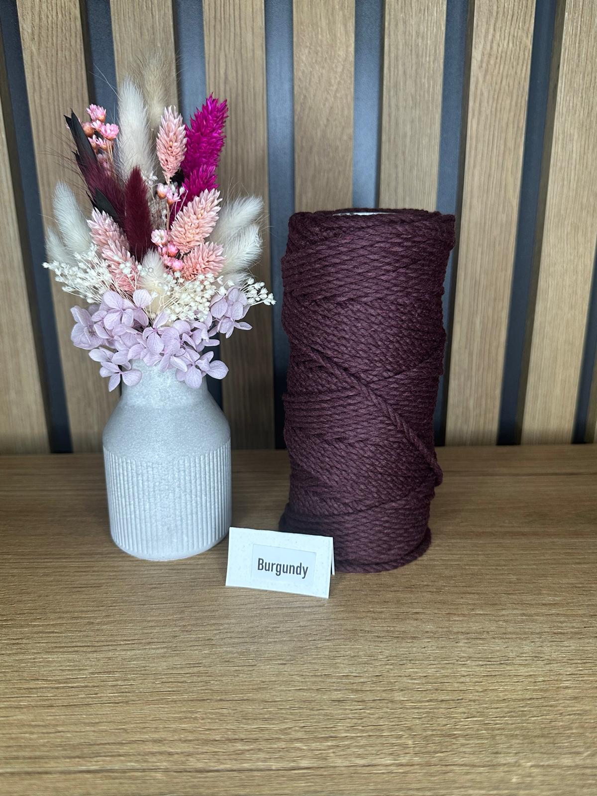 Dunkelrote Garnrolle aus Baumwollkordel in der Farbe Burgundy, dekorativ arrangiert neben einer Vase mit Trockenblumen auf einem Holztisch.
Herzberg Crafts