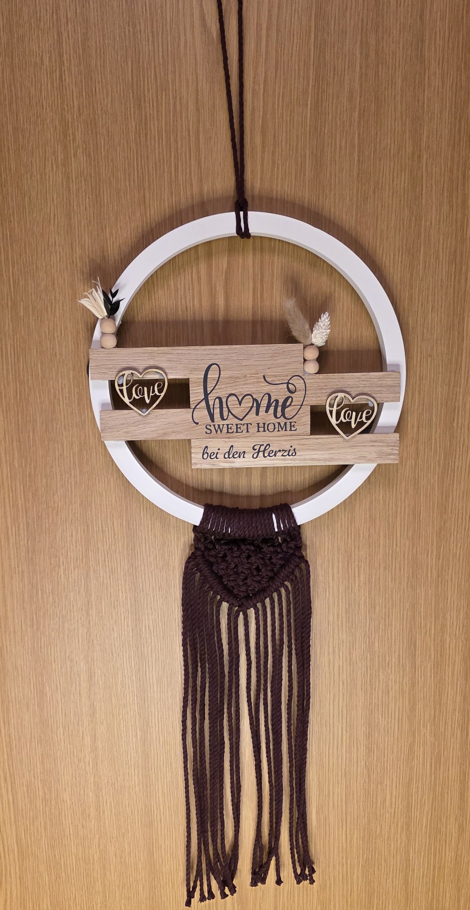 Dekoratives Türschild mit Holzklötzchen die an einem weißem Keracraftring befestigt sind. Mit einem Makramee-Anhänger, Vinyl-Aufschrift ‚home sweet home bei den Herzis‘, verziert mit kleinen Holzherzen und Trockenblumen, aufgehängt an einer Holztür.
Herzberg Crafts