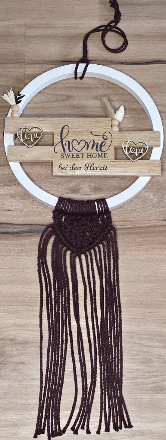 Handgefertigtes Deko-Türschild mit weißem Keracraftring, Holzklötzchen & Vinylschriftzug ‚home sweet home bei den Herzis‘, Burgundy Makramee-Anhänger, liegend fotografiert auf hellem Holzboden.
Herzberg Crafts
